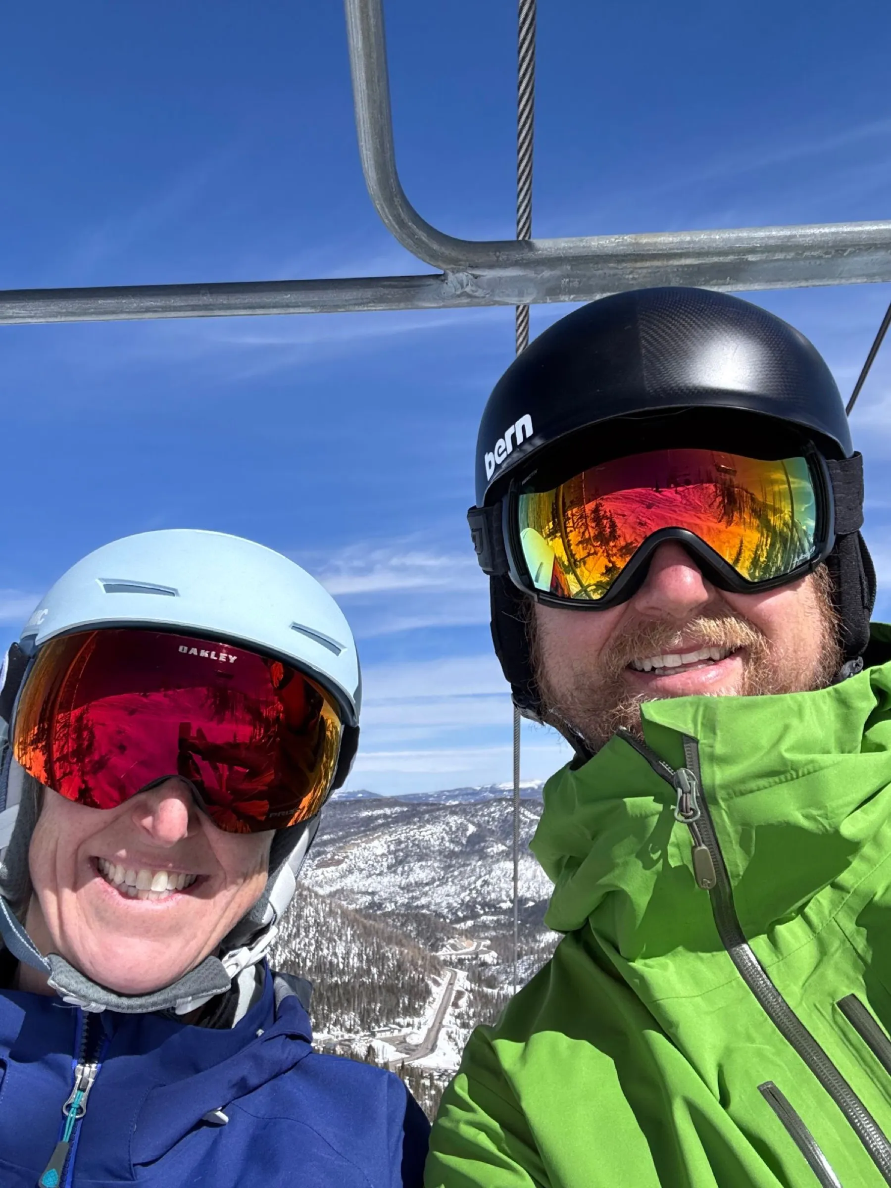 My wife and me smiling on a chairlift in helmets and mirrored goggles, with snowy forested mountains below.