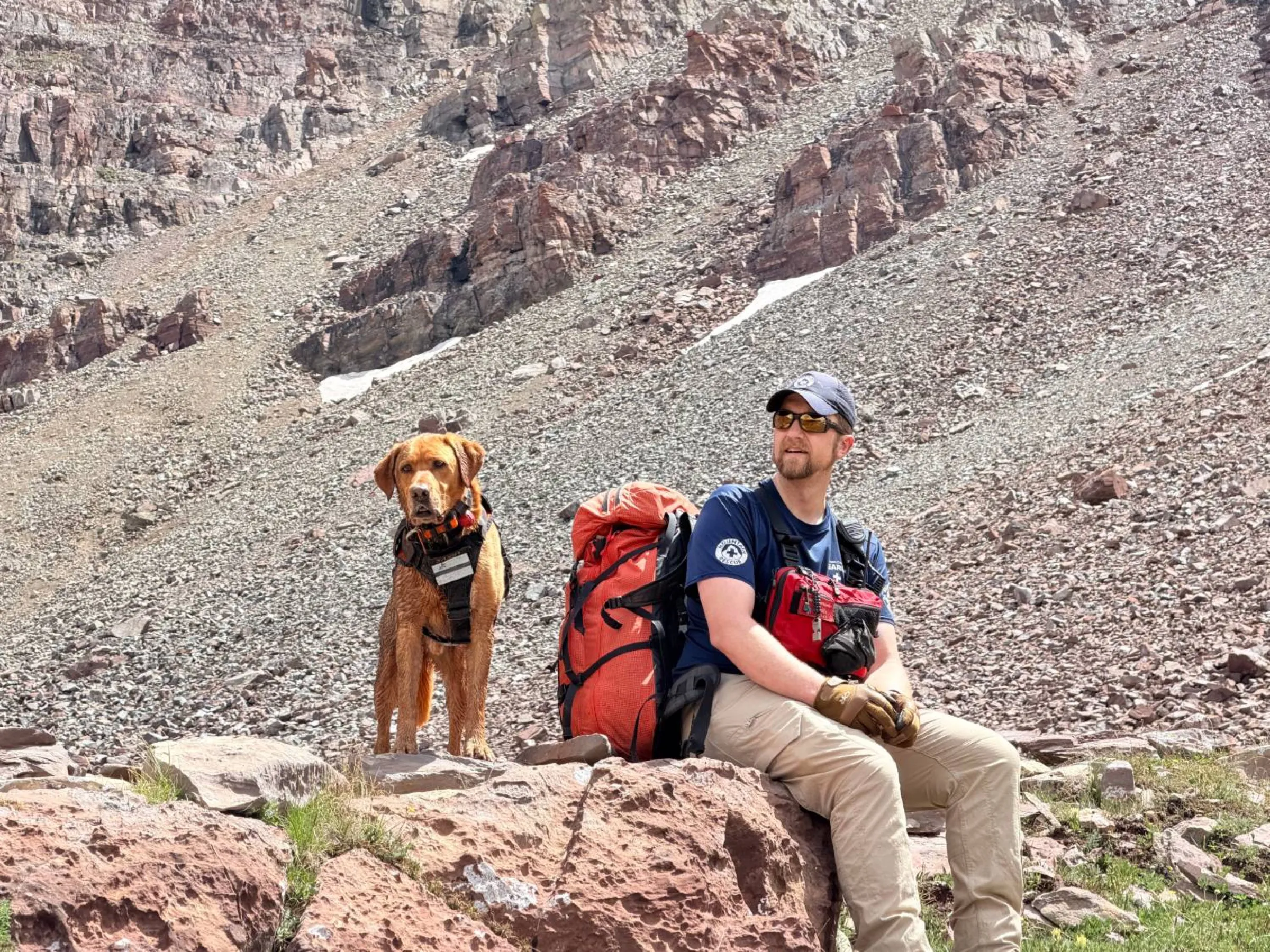 Jay Christianson resting on a rock in search and rescue gear, with Saxby, a red-coated dog in a working harness, on the talus slope behind.
